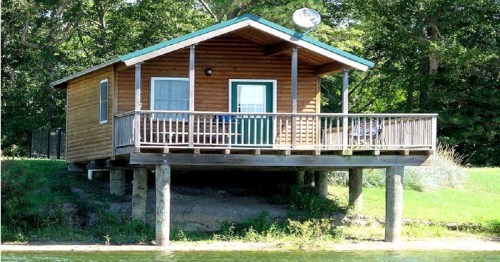 A wooden cabin on stilts by a lake, featuring a porch and satellite dish, surrounded by greenery.