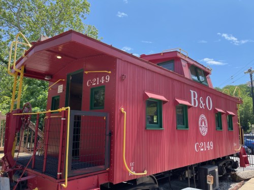 A red caboose labeled "B&O C2149" sits under a clear blue sky, surrounded by trees and a railway.