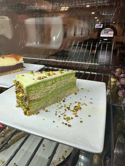 A slice of green pistachio cake on a white plate, displayed in a glass case with other desserts in the background.