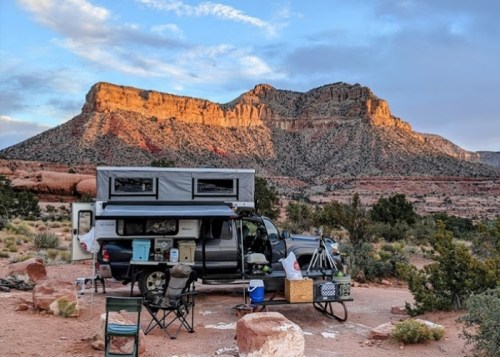 A camper parked in a scenic desert landscape with mountains in the background, surrounded by chairs and outdoor gear.