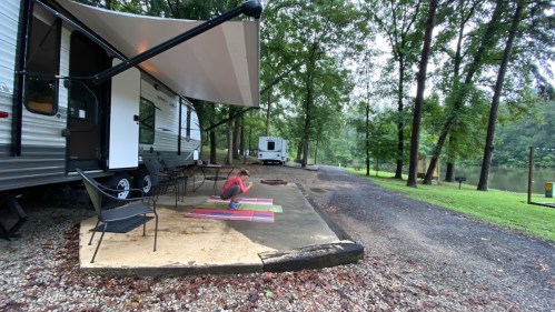 A person practices yoga on mats outside a camper in a wooded campsite by a river.