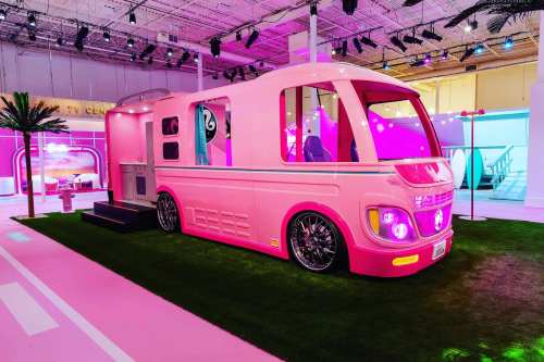 A vibrant pink retro-style camper van displayed in a colorful exhibition space with palm trees and neon lights.
