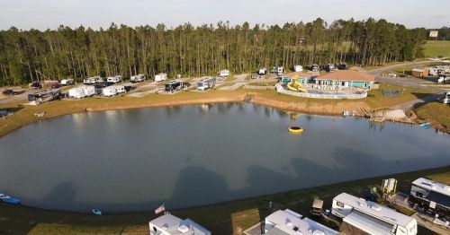 Aerial view of a campground with RVs around a small lake, surrounded by trees and a recreational area.