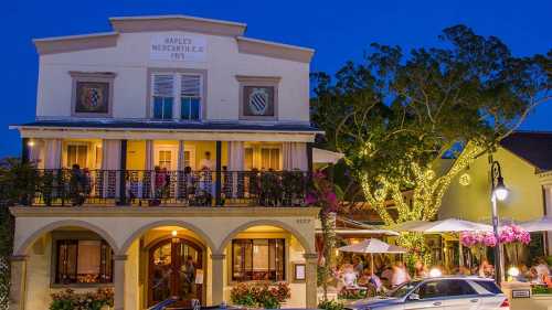 A charming building with outdoor dining, illuminated by lights, surrounded by trees and flowers at dusk.