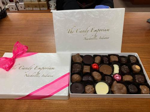 A white box of assorted chocolates from The Candy Emporium, tied with a pink ribbon, on a wooden counter.