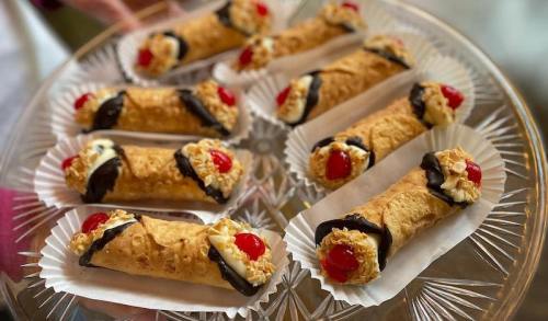 A platter of cannoli filled with cream, topped with cherries and chocolate, arranged on decorative paper liners.