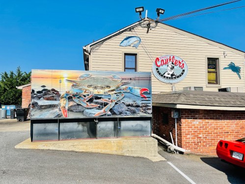 A mural of a crab on a wall outside a seafood restaurant, with a sunset and rocky shoreline in the background.