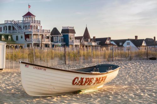 A white boat labeled "Cape May" on sandy beach, with colorful buildings and a wooden fence in the background.