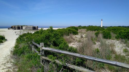 A sandy beach scene with green shrubs, a wooden fence, a historic bunker, and a lighthouse in the distance under a clear blue sky.