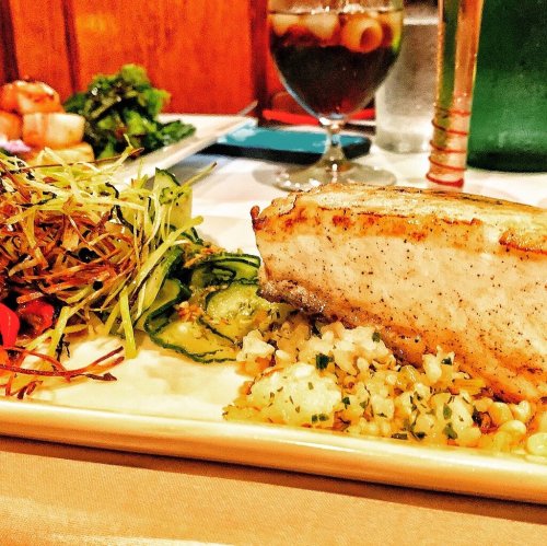 A plated dish featuring grilled fish, vegetables, and a grain side, with a drink and additional food in the background.