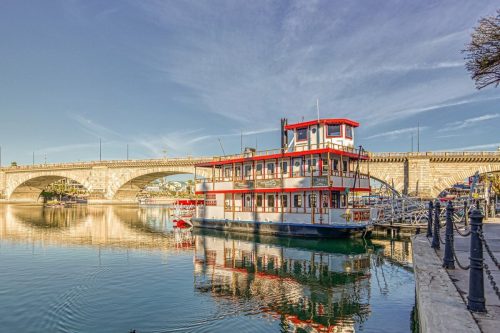 A colorful riverboat docked by a bridge, reflecting in calm waters under a clear blue sky.