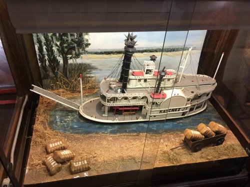 A detailed model of a steamboat displayed in a glass case, with bales of hay and a scenic background.