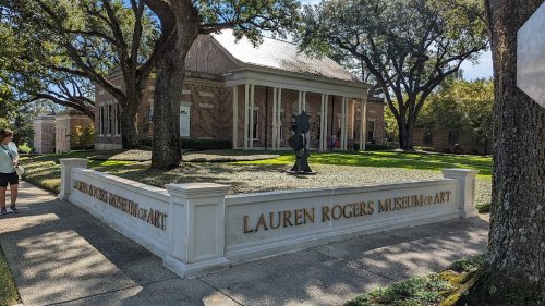 Sign for Lauren Rogers Museum of Art, surrounded by trees and a well-maintained lawn, with visitors in the background.