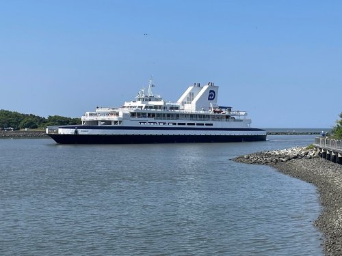 A large ferry boat navigates through calm waters on a clear day, with a rocky shoreline in the foreground.