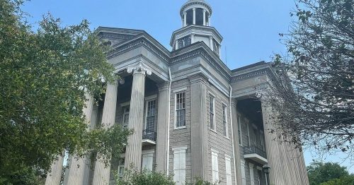 Historic building with tall columns and a dome, surrounded by greenery under a clear blue sky.
