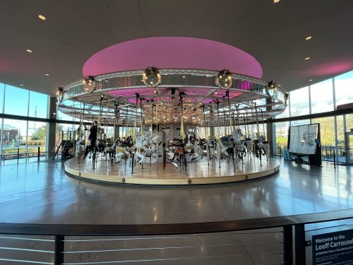 A brightly lit carousel with ornate horses, surrounded by large windows and festive decorations.