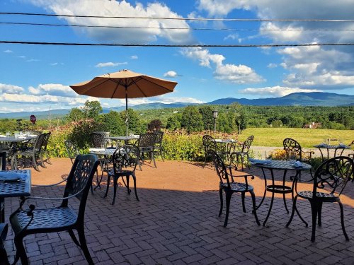 Outdoor dining area with black metal tables and chairs, an umbrella, and a scenic view of mountains and blue skies.