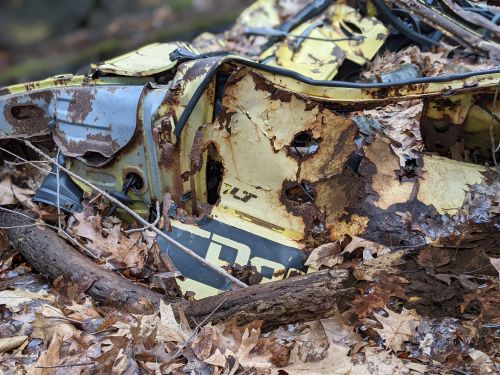 Rusty, weathered metal debris covered in leaves and surrounded by twigs in a forested area.