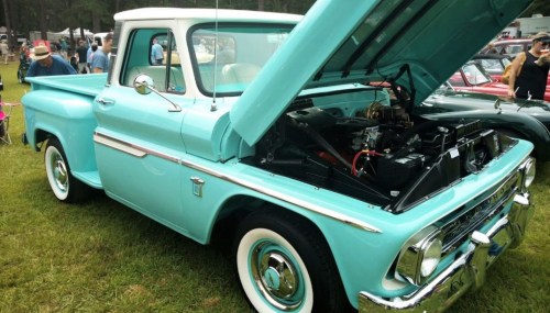 A vintage turquoise pickup truck with the hood open, showcasing its engine, parked at an outdoor event.