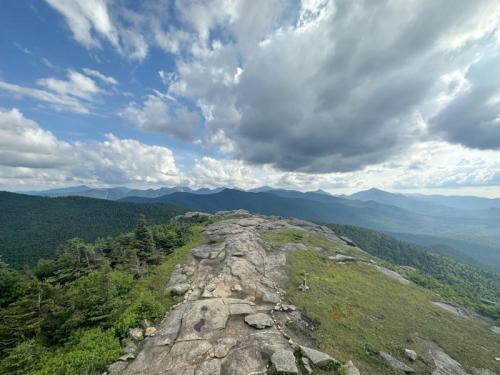 A rocky mountain summit with lush green trees and distant peaks under a cloudy blue sky.