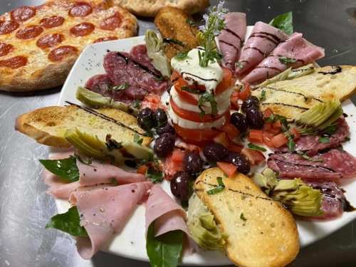 A colorful antipasto platter with meats, cheeses, olives, and vegetables, accompanied by a pepperoni pizza in the background.