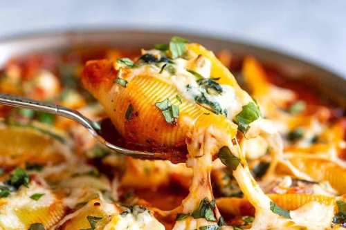 A fork lifting a cheesy stuffed pasta shell from a pan, garnished with fresh herbs in a rich tomato sauce.
