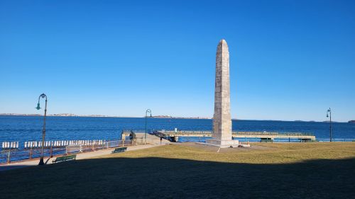 A tall stone obelisk stands near a calm waterfront, with benches and a pier visible under a clear blue sky.