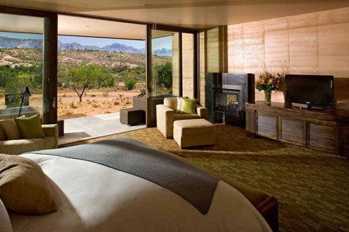 Modern hotel room with large windows, a cozy bed, seating area, and a view of desert landscape and mountains.