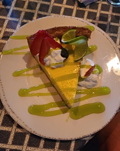 A slice of lemon pie topped with strawberries, lime, and whipped cream, drizzled with green sauce on a white plate.