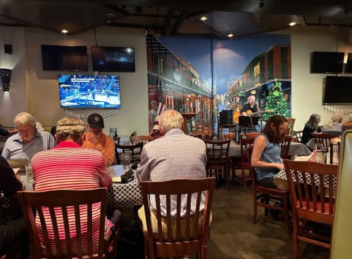 A lively restaurant scene with diners at tables, a musician performing, and a sports game on TV in the background.