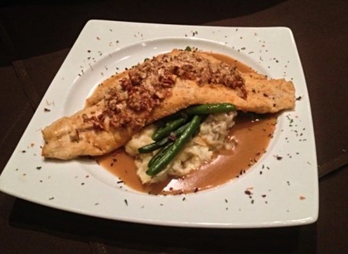 A plated dish featuring a fish fillet topped with nuts, served on mashed potatoes and green beans in a savory sauce.