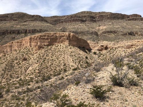 A rugged desert landscape featuring rocky cliffs and sparse vegetation under a cloudy sky.