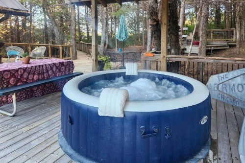 A bubbling hot tub on a wooden deck surrounded by trees, with outdoor furniture and a table nearby.