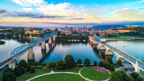 Aerial view of a city skyline with a bridge over a river, surrounded by greenery and a colorful sunset.