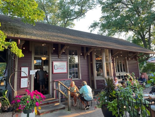 A charming restaurant with a wooden exterior, outdoor seating, and vibrant flowers, set in a lush green environment.