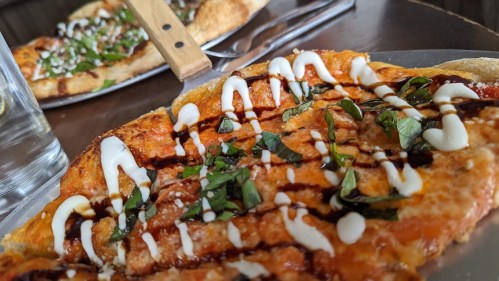 Close-up of a pizza topped with basil, drizzled with sauce and cream, on a metal plate with another pizza in the background.