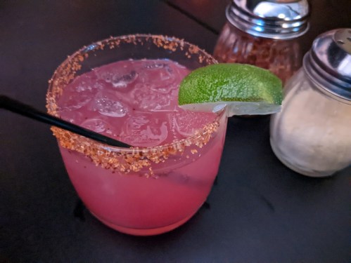 A vibrant pink cocktail with a salted rim, garnished with a lime wedge, sits on a dark table next to condiments.