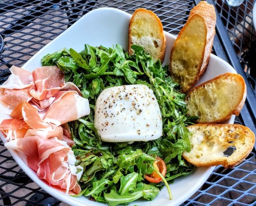 A bowl of arugula topped with a creamy cheese, prosciutto, and toasted bread slices on a black metal table.