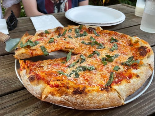 A freshly baked pizza topped with cheese and basil, served on a wooden table with plates in the background.