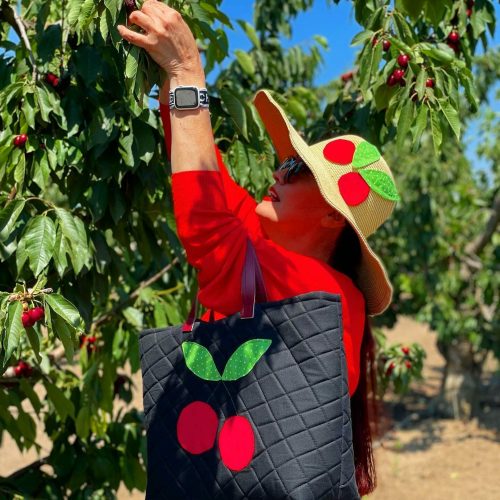 A woman in a red sweater and hat picks cherries from a tree, carrying a black bag with a cherry design.