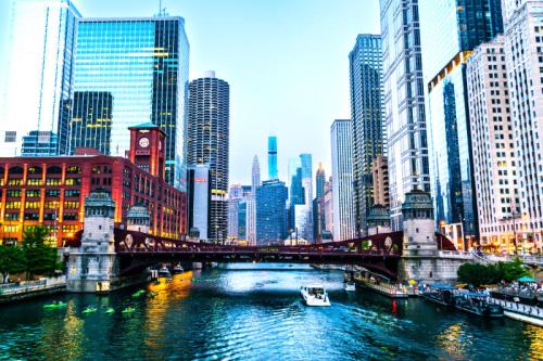 City skyline with skyscrapers along a river, featuring a bridge and boats, under a clear blue sky.