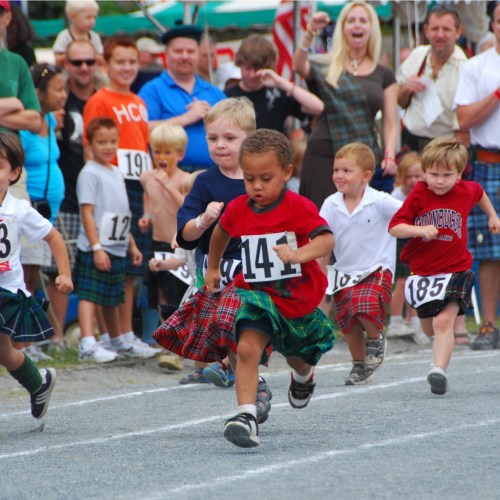 Children in colorful kilts race on a track, with spectators cheering in the background.