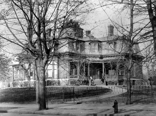 Historic black-and-white photo of a large, ornate house surrounded by trees and a fence, with people on the porch.
