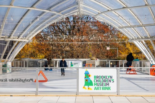 A bright, covered ice rink at the Brooklyn Children's Museum, with people skating and autumn trees in the background.