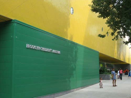 Exterior of the Brooklyn Children's Museum, featuring a bright yellow wall and green entrance, with visitors nearby.