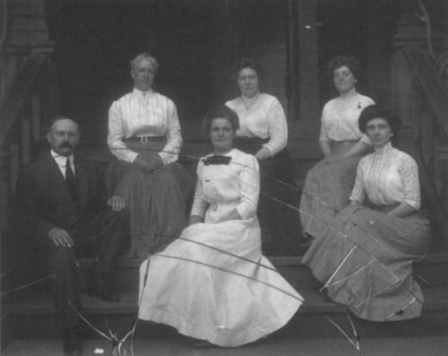 A black and white photo of six women and one man seated on steps, dressed in early 20th-century clothing.