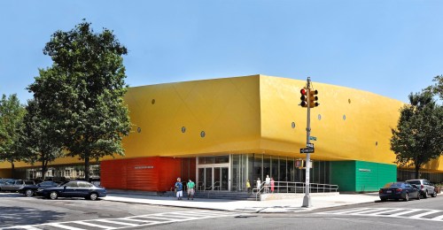 A modern building with a bright yellow exterior and green and orange accents, surrounded by trees and a crosswalk.