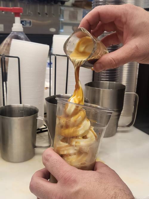A hand pours caramel sauce over a swirl of soft serve ice cream in a clear cup, with metal containers in the background.