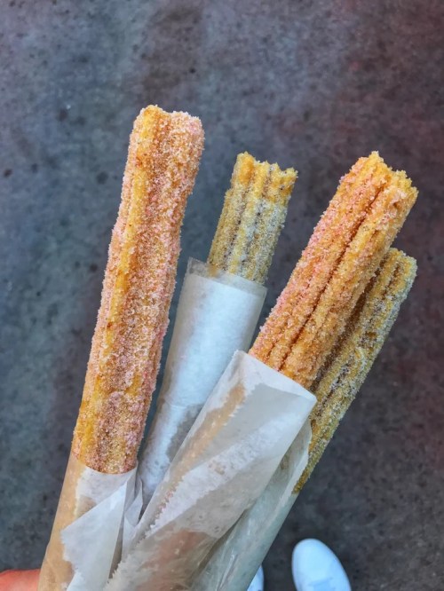 A hand holding three golden churros coated in sugar, wrapped in paper, against a textured background.