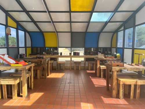 Bright, spacious indoor play area with wooden tables and benches, colorful walls, and various toys scattered around.